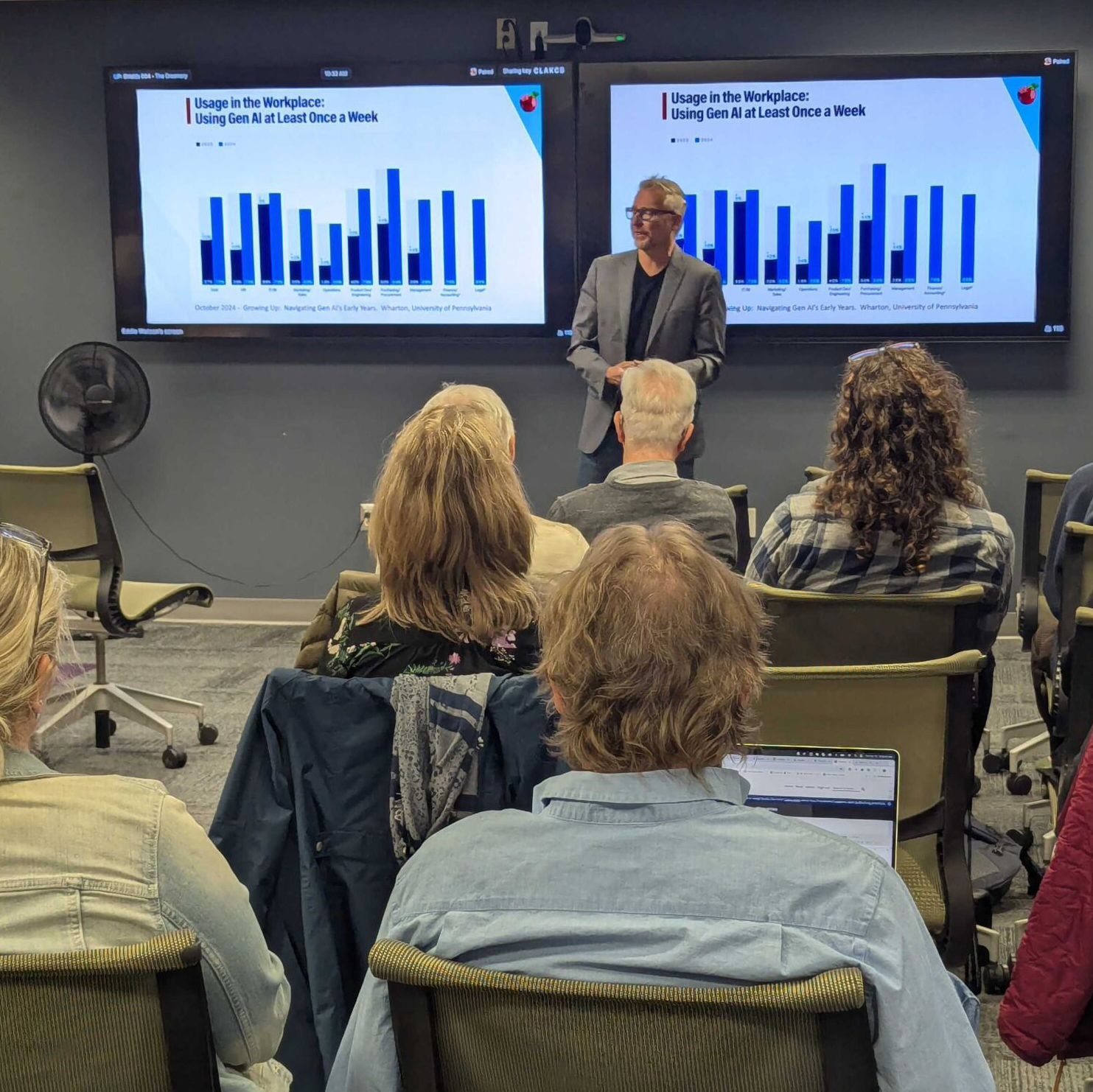 DataInformedCommunity A presenter stands in front of two televisions with bar graphs while a crowd listens
